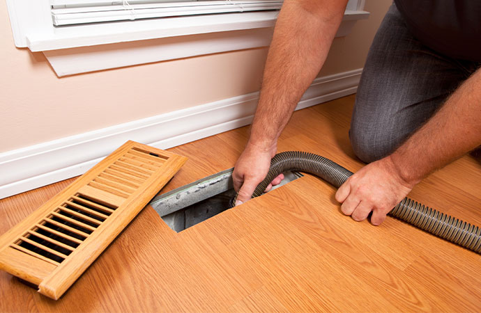 A hand holding a vacuum hose, inserting it into a wooden floor vent for cleaning the air duct